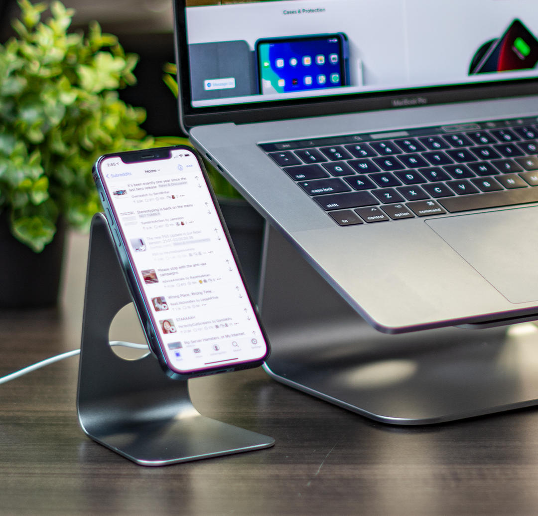 Stand for Apple MagSafe charger crafted from Aluminum and rubber with built-in cable management, charging a iPhone 12 Pro on a desk next to a MacBook Pro on a stand.